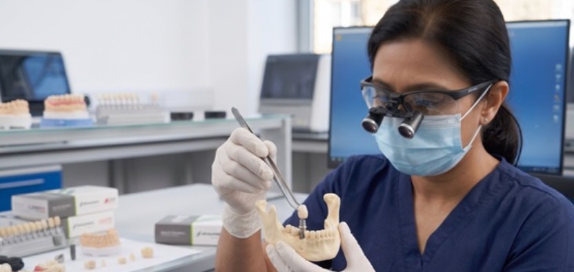 Eine detaillierte, fotorealistische klinische Ansicht eines professionellen Zahnlaboratoriums in Istanbul. Eine weibliche Technikerin überprüft mit Vergrößerungslupen sorgfältig die Mikroprecision eines einzelnen, standardmäßigen, menschlich großen Straumann-Implantat-Abutments. Eine vollständige Standard-Nachbildung eines menschlichen Unterkiefers im Vordergrund bestätigt, dass die Komponenten im realistischen menschlichen Maßstab, klein und nicht überdimensioniert sind, was die technische Strenge der Gewährleistungsbehebung demonstriert.